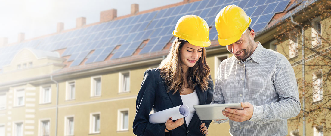 Kobieta z meżczyzną spoglądają w plany remontowe, w tle budynek mieszkalny