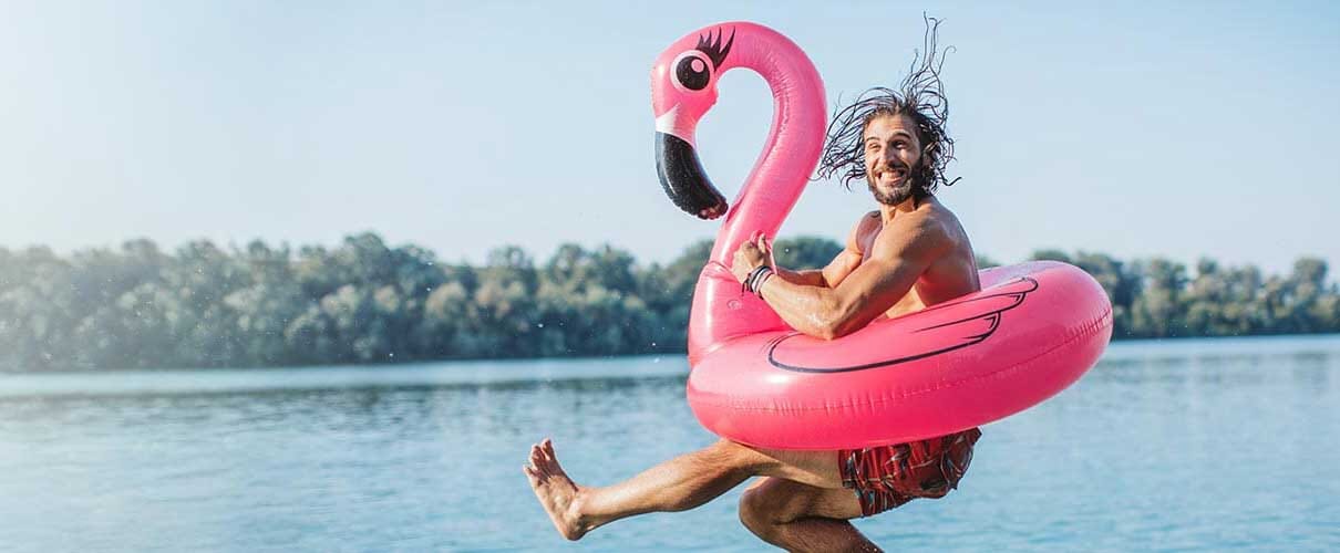 uśmiechnięty mężczyzna skacze do jeziora w kole ratunkowym z różowym flamingiem