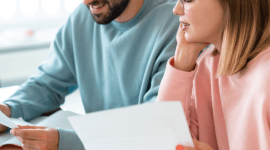 Młoda kobieta w okularach i mężczyzna siedzą przy drewnianym stole, wspólnie przeglądając dokumenty i korzystając z laptopa w jasnym wnętrzu.