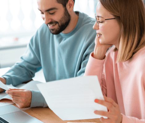 Młoda kobieta w okularach i mężczyzna siedzą przy drewnianym stole, wspólnie przeglądając dokumenty i korzystając z laptopa w jasnym wnętrzu.