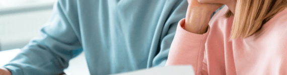 Młoda kobieta w okularach i mężczyzna siedzą przy drewnianym stole, wspólnie przeglądając dokumenty i korzystając z laptopa w jasnym wnętrzu.