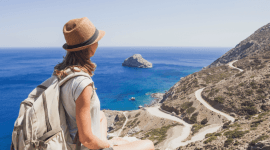 Podróżniczka w słomkowym kapeluszu i z plecakiem siedzi na skalistym szczycie, patrząc na błękitne morze, małą wysepkę oraz krętą drogę wijącą się wzdłuż górzystego wybrzeża w słoneczny dzień.