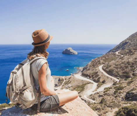 Podróżniczka w słomkowym kapeluszu i z plecakiem siedzi na skalistym szczycie, patrząc na błękitne morze, małą wysepkę oraz krętą drogę wijącą się wzdłuż górzystego wybrzeża w słoneczny dzień.