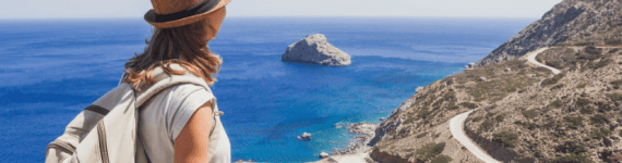 Podróżniczka w słomkowym kapeluszu i z plecakiem siedzi na skalistym szczycie, patrząc na błękitne morze, małą wysepkę oraz krętą drogę wijącą się wzdłuż górzystego wybrzeża w słoneczny dzień.