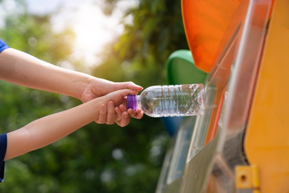 Dwie ręce wkładają plastikową butelkę do śmietnika na plastiki.